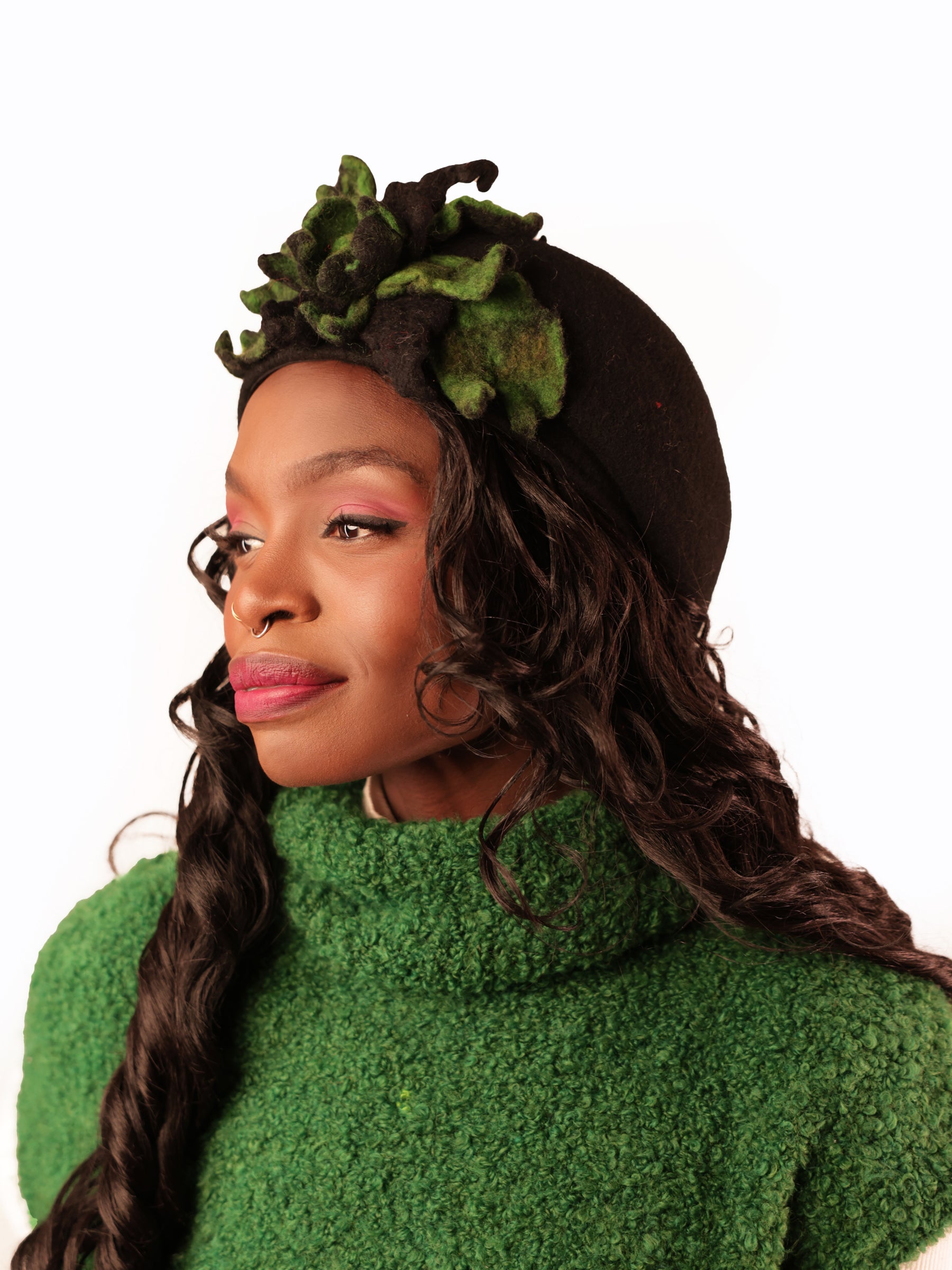 Side profile of black wool beret with sculptural green felt floral embellishment, model looking fully to the right.