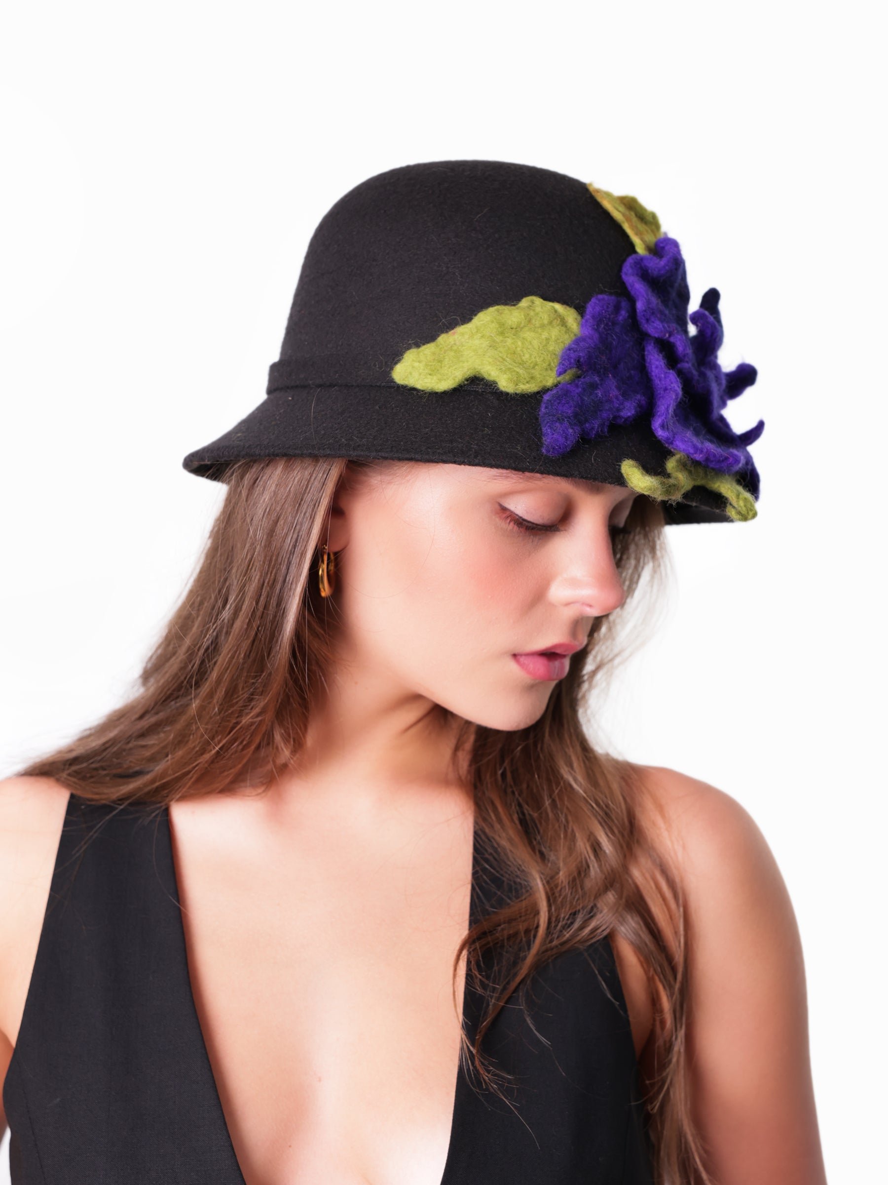 Side view of a black cloche hat featuring sculptural violet felt flowers and green leaf accents.