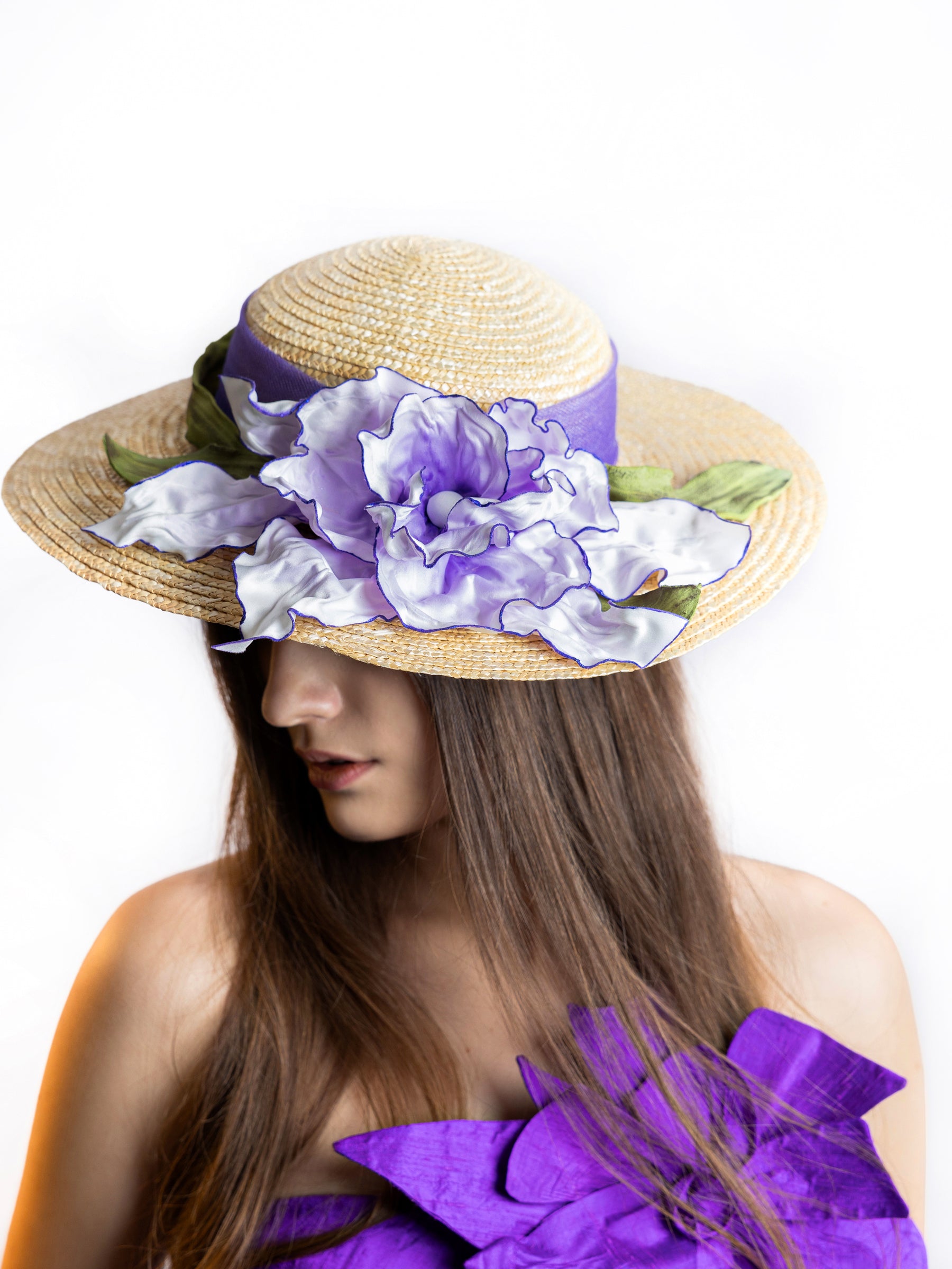 Model wearing a straw summer hat with satin floral embellishments and purple ribbon, angled view.